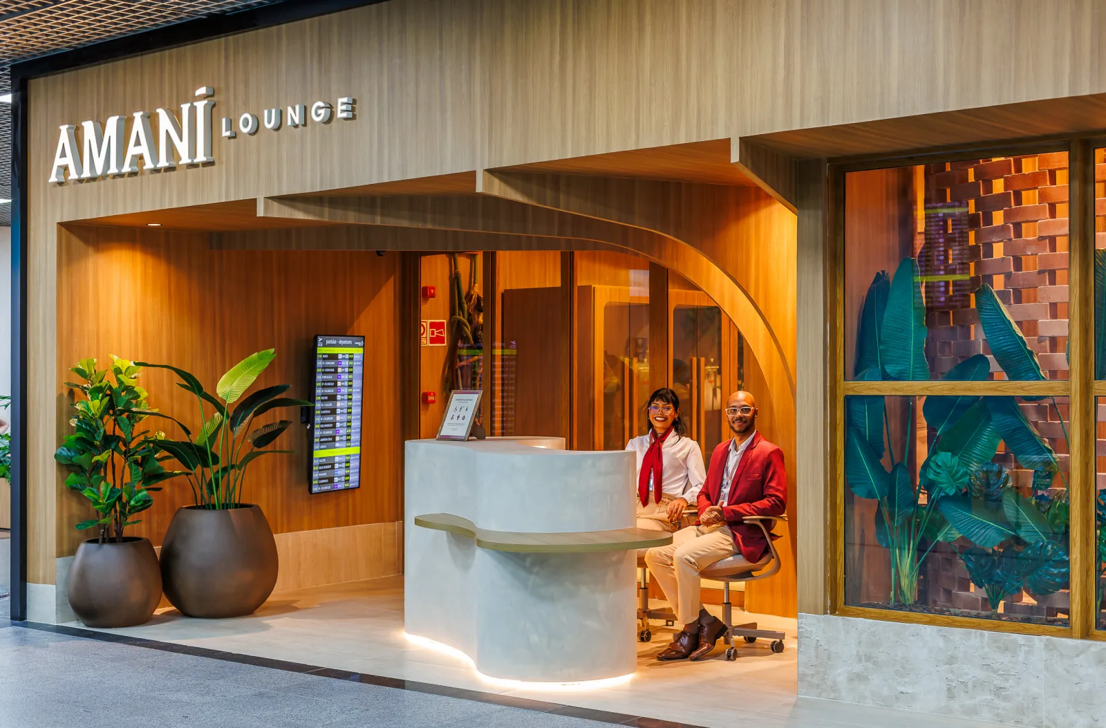 Entrada do Amaní Lounge, com fachada de madeira e dois atendentes de uniforme vermelho sentados no balcão da recepção